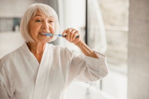 Mature woman with white hair wearing a white bathrobe brushing her teeth with a blue toothbrush