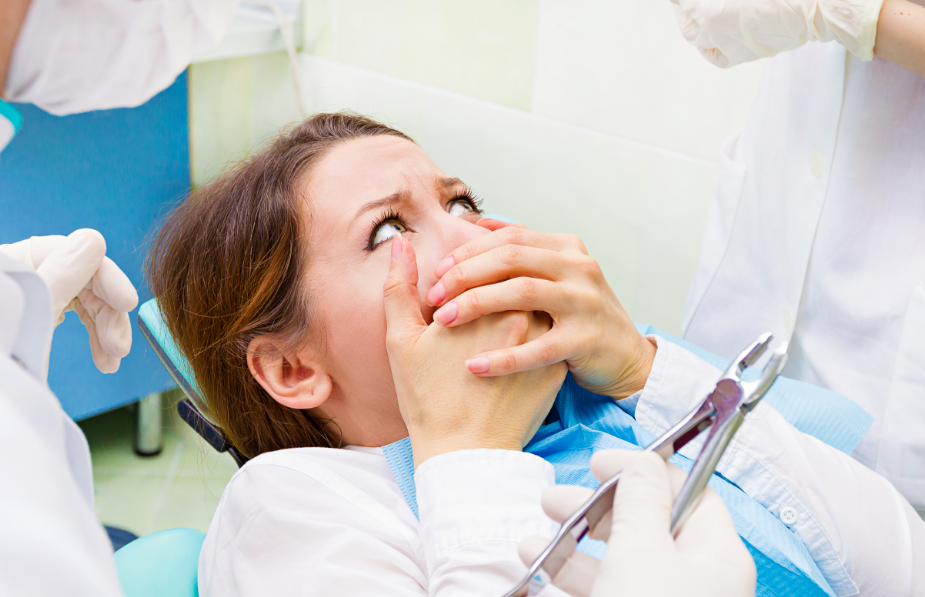 Woman in dental chair covering mouth and looking terrified while dentist hands hover over her.